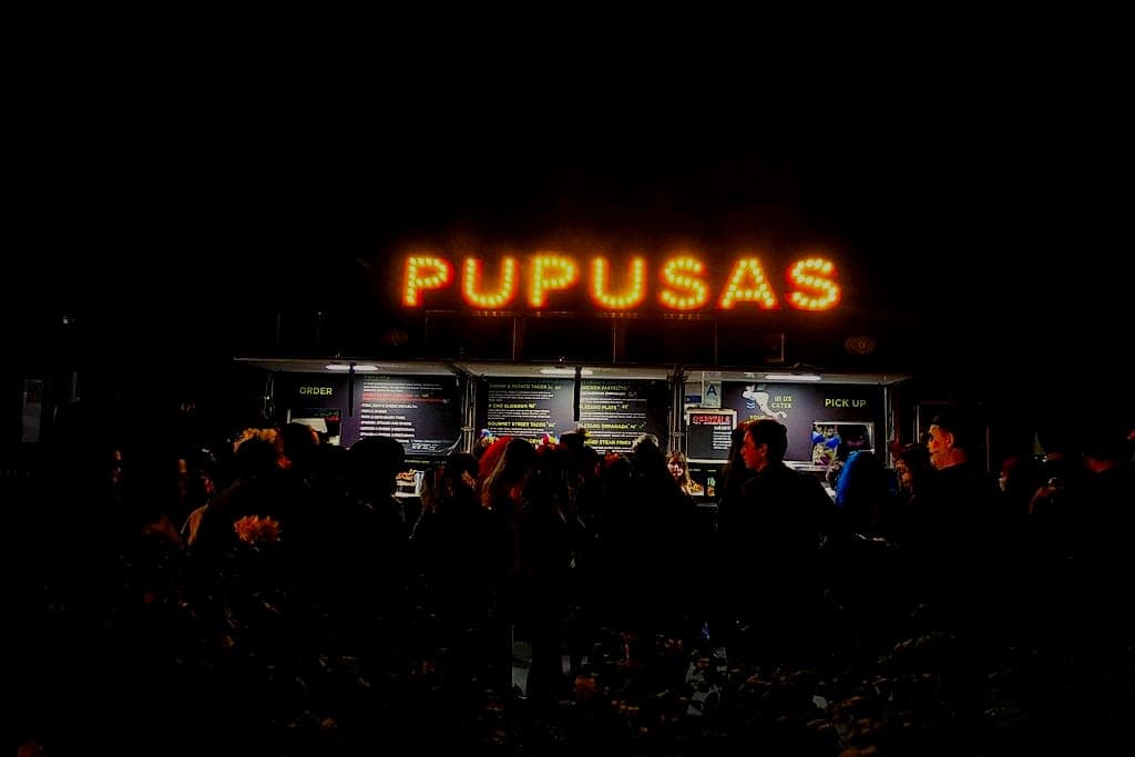 A neon sign reading PUPUSAS above a food stall at night